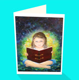 A young girl sitting with her legs crossed reading a large book in her lap. Watch the glow and luminescence coming from the book. By Australian artist Emma Harris and published as a greeting card by Cloud Publishing
