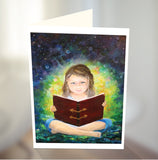 A young girl sitting with her legs crossed reading a large book in her lap. Watch the glow and luminescence coming from the book. By Australian artist Emma Harris and published as a greeting card by Cloud Publishing